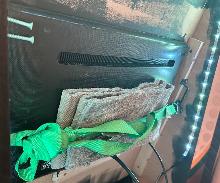 The rear of the monitor resting on a 1x2 ledge, secured with side screws and a strap across the back